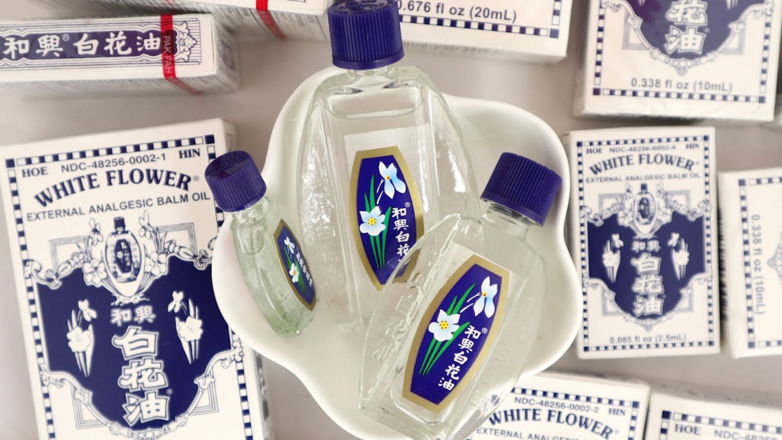 Three different sized bottles of White Flower oil in small dish surrounded by boxes of White Flower oil